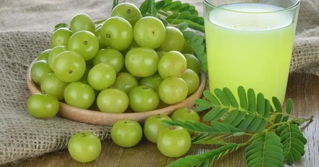 A bowl of fresh green berries beside a glass of pale green juice, with green leaves scattered on a textured wooden surface.