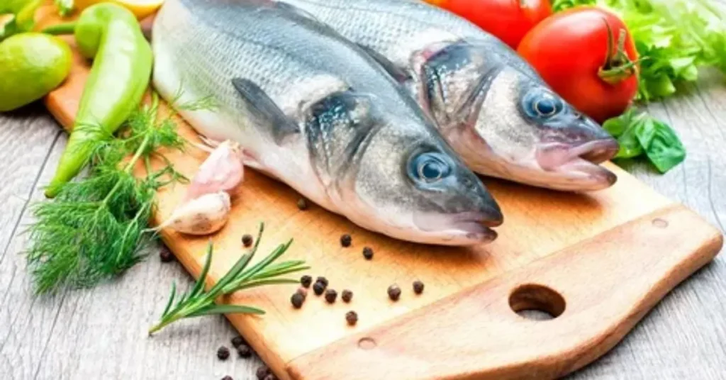 Two fresh fish on a wooden cutting board surrounded by vegetables, including tomatoes, peppers, garlic, and herbs.