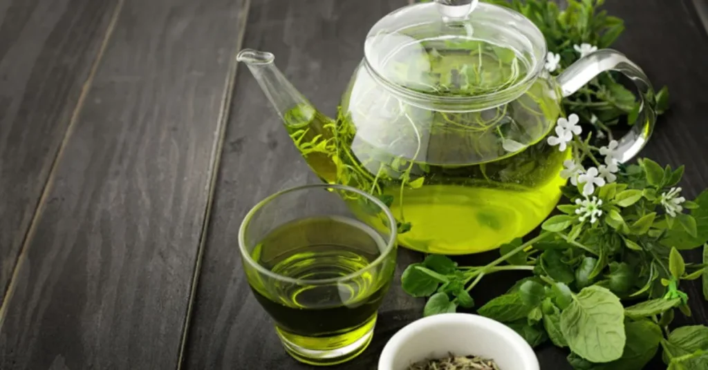 A glass teapot filled with green herbal tea, accompanied by a glass and a small bowl of loose herbs on a wooden surface.
