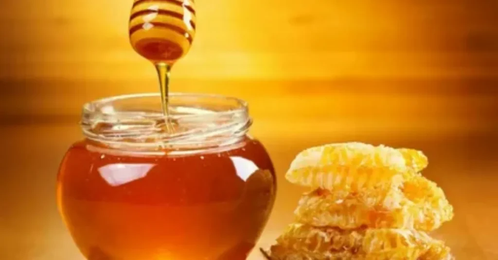 A jar of golden honey with a honey dipper drizzling honey, next to a stack of honeycomb, against a warm, blurred background.
