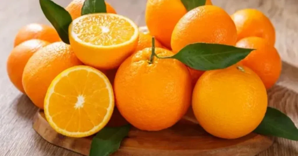 A pile of bright orange oranges with green leaves, featuring one halved orange showcasing its juicy interior on a wooden surface.