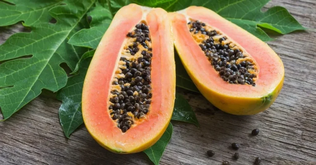 A fresh, halved papaya reveals its vibrant orange flesh and black seeds, resting on green papaya leaves against a wooden surface.