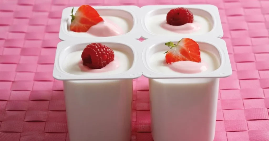 Four creamy yogurt cups topped with fresh strawberries and raspberries, placed on a vibrant pink woven surface.