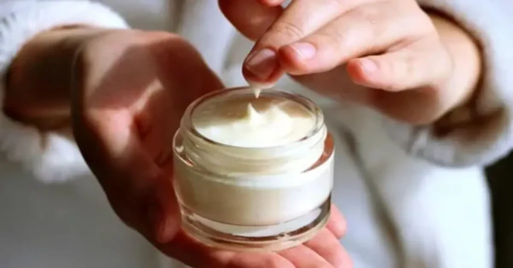 A detailed image shows a person submerging their fingernail into a jar containing soft cream-white facial moisturizer. The person uses one hand to hold the jar while selecting a small portion of the cream with their other hand during skincare application.