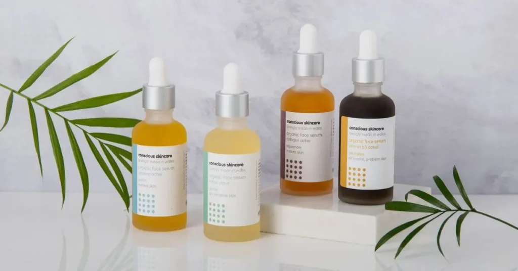 A display of four dropper bottles of organic face serums from Conscious Skincare, each with different colored labels and liquid contents, arranged on a white surface with green palm leaves as decoration, against a light marble background.